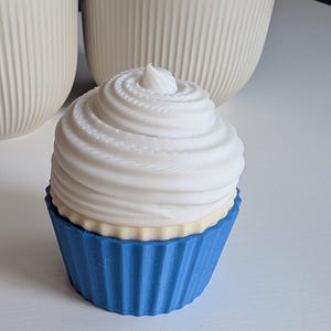 May include: A decorative cupcake with a white swirl frosting and a blue cupcake liner. The cupcake has a light yellow layer between the frosting and the liner. The background includes two white, ribbed vases.