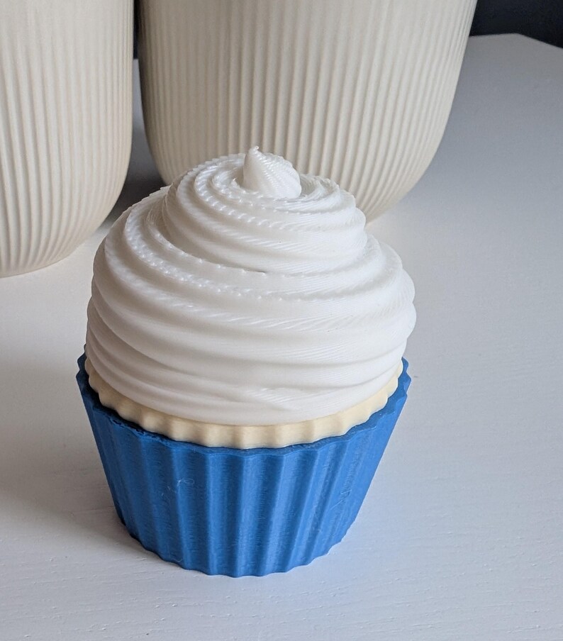 May include: A decorative cupcake with a white swirl frosting and a blue cupcake liner. The cupcake has a light yellow layer between the frosting and the liner. The background includes two white, ribbed vases.