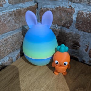 K&ouml;nnte beinhalten: Ein Objekt in Form eines Hasen mit Farbverlauf, mit langen Ohren und einer kleinen orangefarbenen Karottenfigur mit gr&uuml;nem Oberteil. Der Hase ist blau, gr&uuml;n und lila. Die Karotte hat ein l&auml;chelndes Gesicht.
