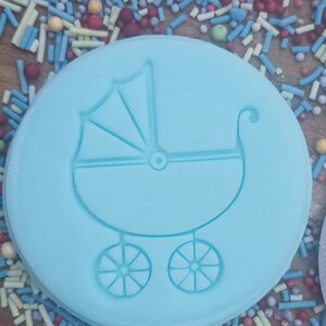 May include: A light blue, circular mold featuring a baby carriage design. The carriage has a canopy, wheels, and a curved handle. The mold is surrounded by colorful sprinkles, suggesting a theme of celebration or a baby shower.