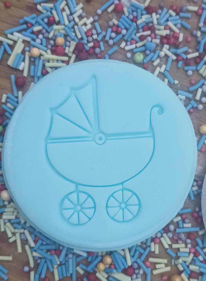 May include: A light blue, circular mold featuring a baby carriage design. The carriage has a canopy, wheels, and a curved handle. The mold is surrounded by colorful sprinkles, suggesting a theme of celebration or a baby shower.