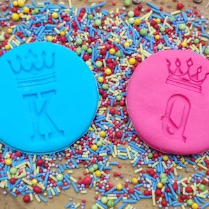 May include: Two round, colorful cookie stamps with crown designs and the letters "K" and "Q" are surrounded by colorful sprinkles. The blue stamp has a "K" and the pink stamp has a "Q". The background is a wooden surface.