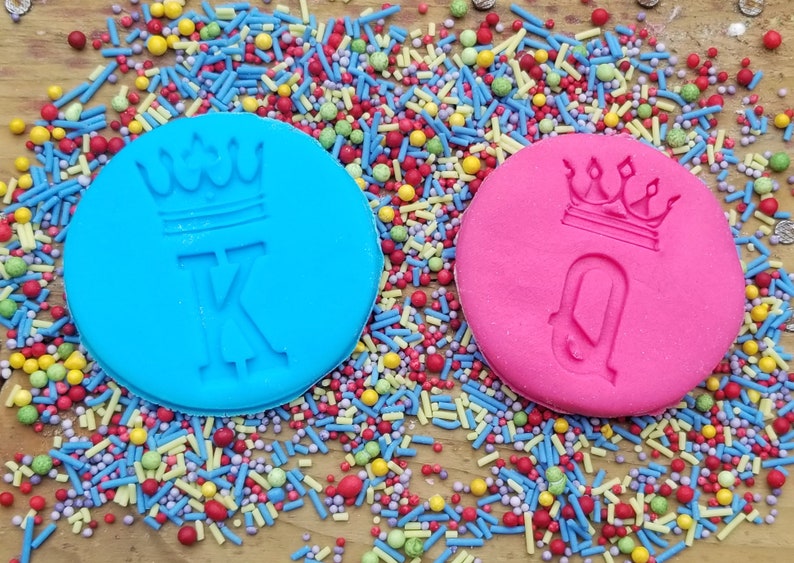 May include: Two round, colorful cookie stamps with crown designs and the letters "K" and "Q" are surrounded by colorful sprinkles. The blue stamp has a "K" and the pink stamp has a "Q". The background is a wooden surface.