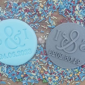 May include: Two round, blue and gray cookie cutters with the letters "S&I" and "I&S" and the date "24.06.2023" on them. The cutters are surrounded by colorful sprinkles.