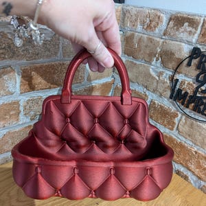 May include: A red decorative handbag-shaped container with a quilted pattern. The container has a handle and a rounded base. The color is a deep, rich red. The container is designed to hold small items.