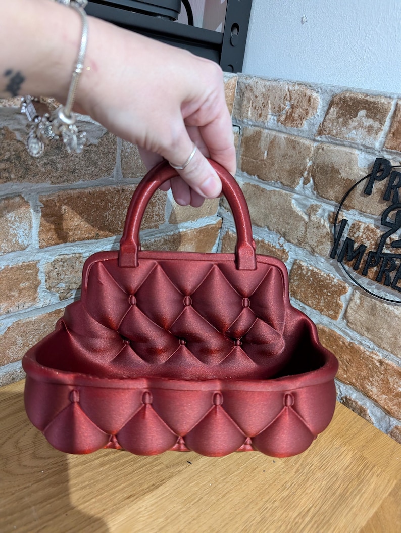 May include: A red decorative handbag-shaped container with a quilted pattern. The container has a handle and a rounded base. The color is a deep, rich red. The container is designed to hold small items.