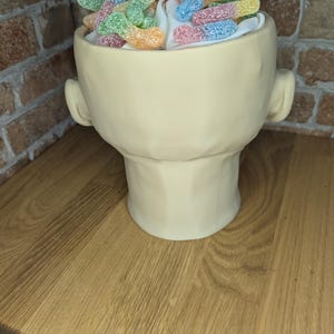 May include: A cream-colored, head-shaped ceramic container filled with white fluff and colorful gummy candies. The container has ear-like protrusions on either side. The candies are in various colors, including green, yellow, pink, and blue.