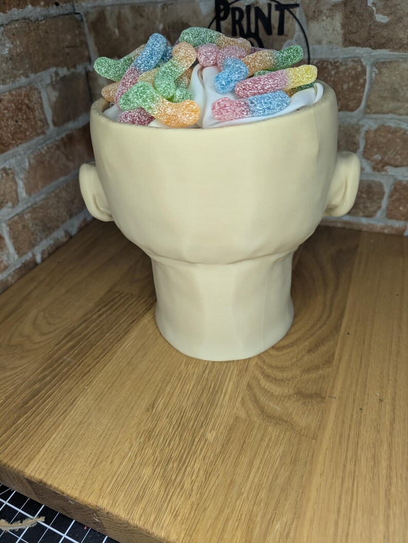 May include: A cream-colored, head-shaped ceramic container filled with white fluff and colorful gummy candies. The container has ear-like protrusions on either side. The candies are in various colors, including green, yellow, pink, and blue.