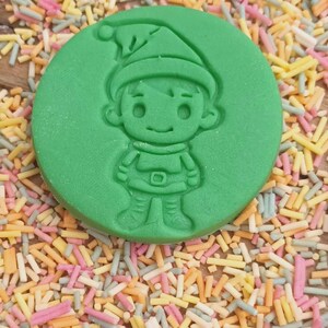 May include: Green plastic cookie cutter shaped like a cartoon elf wearing a red hat. The elf is smiling and has a belt around its waist.