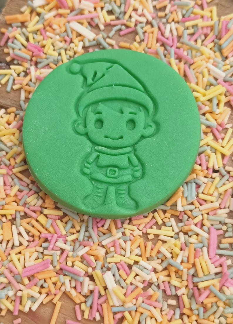 May include: Green plastic cookie cutter shaped like a cartoon elf wearing a red hat. The elf is smiling and has a belt around its waist.