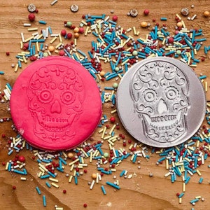 Puede incluir: Dos adornos redondos con dise&ntilde;os de calaveras de az&uacute;car. Uno es rosa brillante, el otro plateado. Ambos est&aacute;n rodeados de chispas de colores sobre una superficie de madera. La calavera de az&uacute;car es un motivo del D&iacute;a de Muertos.