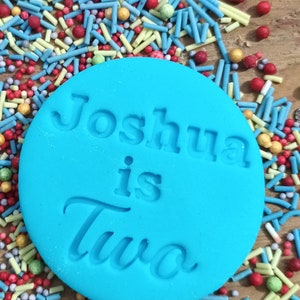 May include: A turquoise fondant circle with the embossed text "Joshua is Two" sits atop a bed of colorful sprinkles. The sprinkles include blue, red, yellow, and green shapes and spheres, creating a festive, celebratory theme.
