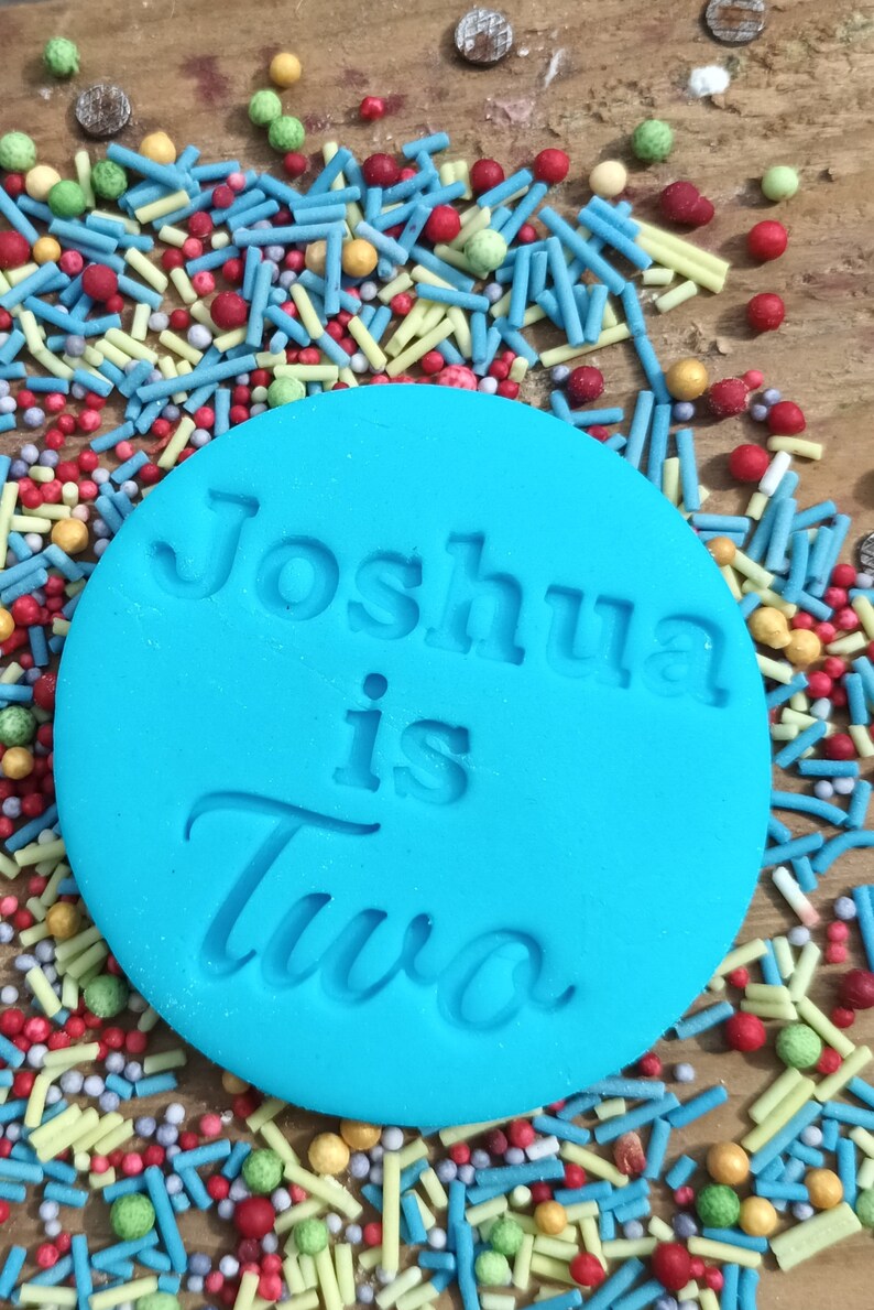 May include: A turquoise fondant circle with the embossed text "Joshua is Two" sits atop a bed of colorful sprinkles. The sprinkles include blue, red, yellow, and green shapes and spheres, creating a festive, celebratory theme.