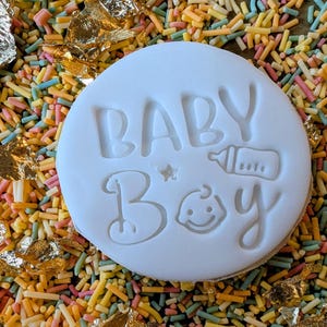 May include: A white, circular cookie with the words "BABY BOY" embossed on its surface, along with a baby bottle and a smiling baby face. The cookie is surrounded by colorful sprinkles and gold leaf.