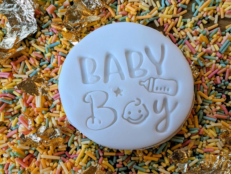 May include: A white, circular cookie with the words "BABY BOY" embossed on its surface, along with a baby bottle and a smiling baby face. The cookie is surrounded by colorful sprinkles and gold leaf.