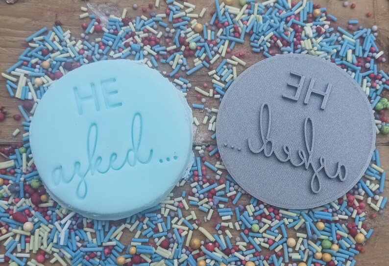 May include: Two round, blue and gray cookie cutters with the text "HE asked..." and "...he said" on them. The cookie cutters are surrounded by colorful sprinkles.