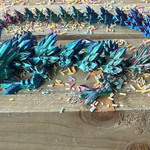 May include: A 3D printed iridescent dragon figurine with a teal, blue, and purple color gradient. The dragon is curled up and has a spiky, crystal-like texture. The dragon is lying on a wooden surface with colorful sprinkles scattered around it.