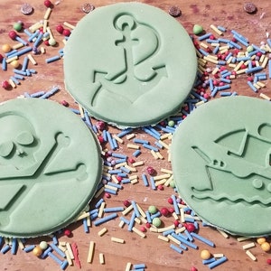 May include: Three round, light green pirate-themed decorations with embossed designs. One features a skull and crossbones, another an anchor, and the third a ship. Colorful sprinkles and a wooden surface complete the scene.