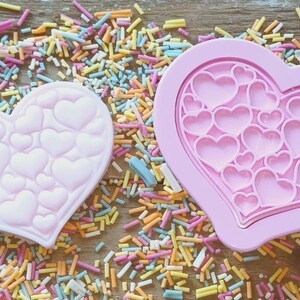 Puede incluir: Dos moldes rosas en forma de coraz&oacute;n, uno con un patr&oacute;n de coraz&oacute;n en relieve y otro con recortes de coraz&oacute;n. Los moldes est&aacute;n rodeados de chispas de colores, lo que sugiere un tema festivo, ideal para hornear o manualidades.