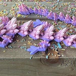 May include: A pink and purple crystal dragon figurine with a gradient color scheme. The dragon is made of plastic and has a detailed design. The dragon is lying on a wooden surface with colorful sprinkles scattered around it.