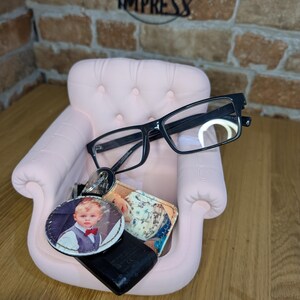 May include: A small, pink decorative chair holds black-framed eyeglasses, a keychain with a photo of a child, and a small wallet. The chair is set against a brick wall with a metal sign that reads "2 Impress".