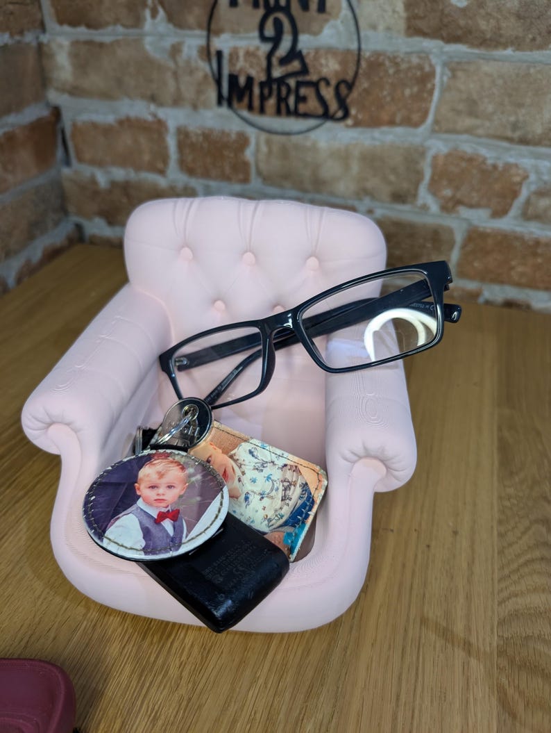 May include: A small, pink decorative chair holds black-framed eyeglasses, a keychain with a photo of a child, and a small wallet. The chair is set against a brick wall with a metal sign that reads "2 Impress".
