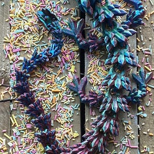 May include: A 3D printed iridescent blue and purple dragon figurine with a spiky back and clawed feet. The dragon is lying on a wooden surface covered in colorful sprinkles.