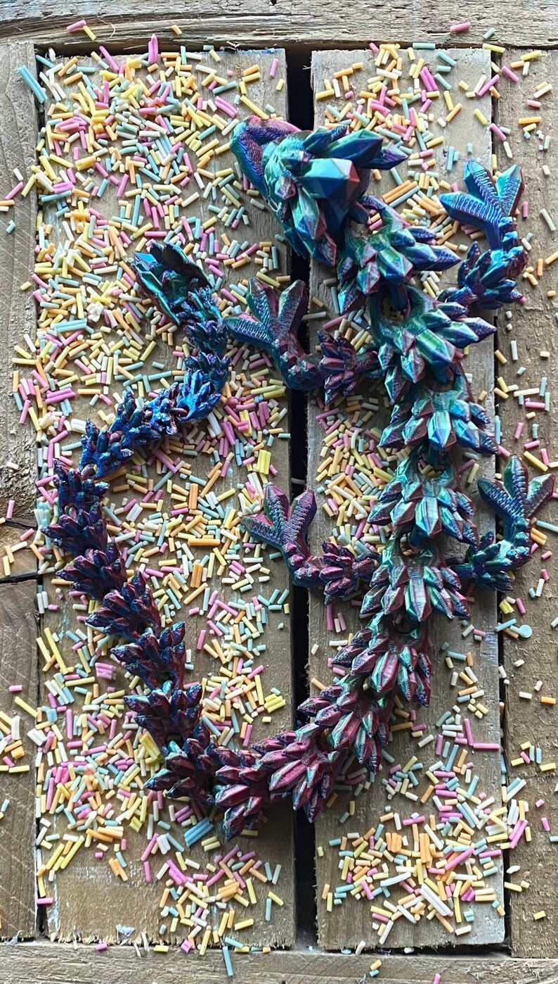 May include: A 3D printed iridescent blue and purple dragon figurine with a spiky back and clawed feet. The dragon is lying on a wooden surface covered in colorful sprinkles.