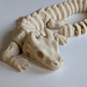 May include: A white, articulated, skeletal dragon figurine with a long tail and a detailed head. The dragon is posed in a crawling position.