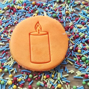 May include: A round, orange biscuit featuring a candle design. The biscuit is set against a backdrop of colourful sprinkles in shades of blue, yellow, red, and green. The sprinkles are scattered on a wooden surface, creating a vibrant display.