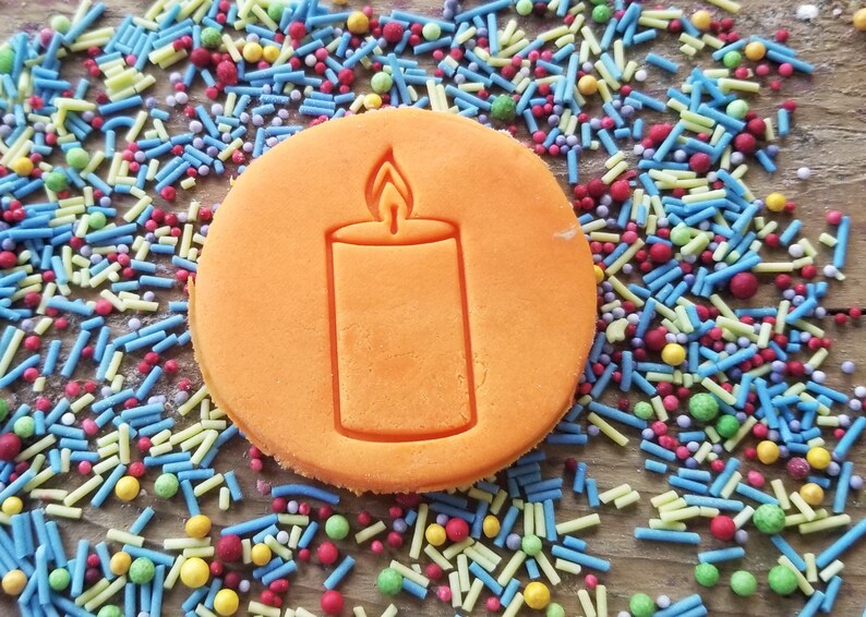 May include: A round, orange biscuit featuring a candle design. The biscuit is set against a backdrop of colourful sprinkles in shades of blue, yellow, red, and green. The sprinkles are scattered on a wooden surface, creating a vibrant display.