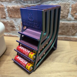 May include: A multi-colored battery holder with the word "BATTERY" and a recycling symbol. The holder contains two red and yellow AA batteries. The holder is made of metal and has a unique design.