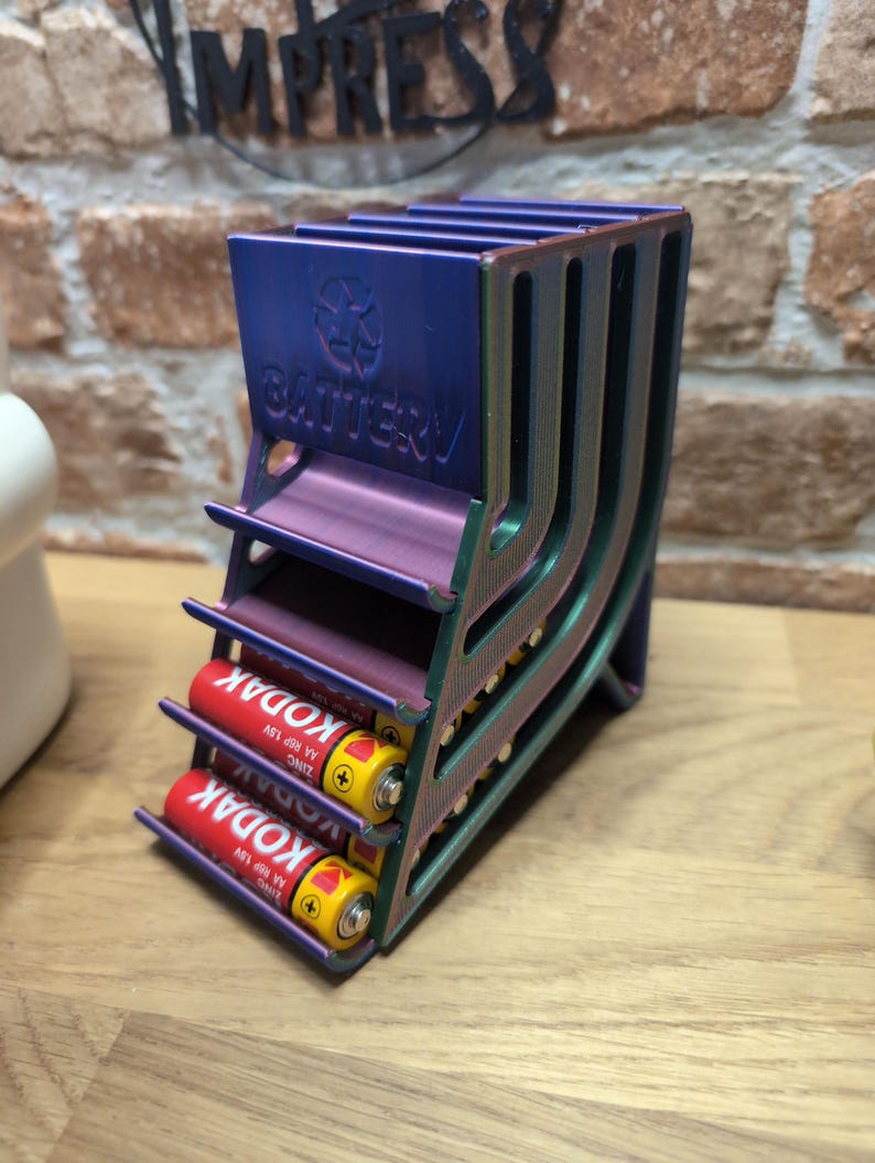 May include: A multi-colored battery holder with the word "BATTERY" and a recycling symbol. The holder contains two red and yellow AA batteries. The holder is made of metal and has a unique design.