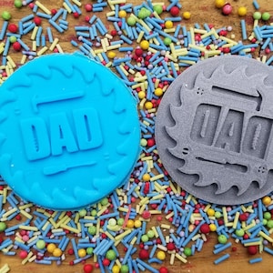 May include: Two round cookie stamps with the word "DAD" in a bold font. One stamp is light blue, the other is gray. Both are surrounded by a decorative border. The stamps are on a wooden surface covered in colorful sprinkles.
