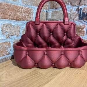 May include: A burgundy-colored, handbag-shaped decorative container with a handle. The container has a quilted pattern on the front and back, with a rounded base. The handle is also burgundy. The container is sitting on a wooden surface.