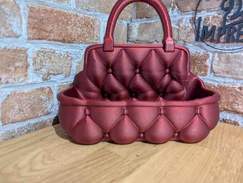 May include: A burgundy-colored, handbag-shaped decorative container with a handle. The container has a quilted pattern on the front and back, with a rounded base. The handle is also burgundy. The container is sitting on a wooden surface.