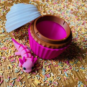 May include: A pink cupcake-shaped container with a brown rim and a white, swirled lid. A pink axolotl figurine rests beside the container, surrounded by colorful sprinkles. The container is open, revealing a bright pink interior.