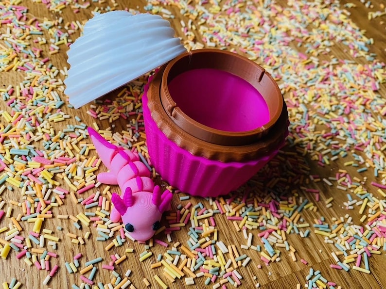 May include: A pink cupcake-shaped container with a brown rim and a white, swirled lid. A pink axolotl figurine rests beside the container, surrounded by colorful sprinkles. The container is open, revealing a bright pink interior.