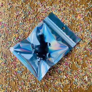 May include: A black and white fox figurine inside a holographic zip-lock bag. The bag is on a wooden surface covered in colorful sprinkles. The fox has white markings around its eyes and chest.