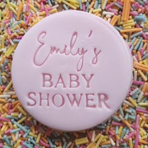 Puede incluir: Una galleta redonda rosa con el texto "Emily's Baby Shower" en letras rosas.