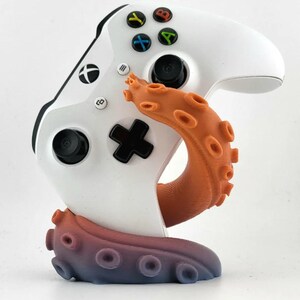 May include: A white Xbox controller is held by an orange and blue tentacle-shaped stand. The controller has black buttons and coloured action buttons. The stand is a gradient of orange to blue.