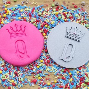 May include: A close-up shot features two circular objects with a crown and the letter "Q" embossed on them. One is pink, the other is gray. Colorful sprinkles surround the objects, creating a festive and playful scene. The objects are likely for baking or crafting.