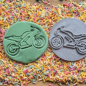 May include: Two circular motorcycle-themed cookie cutters. One is green, the other is gray. Both have a motorcycle design. The cutters are surrounded by colorful sprinkles.