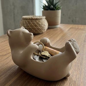 K&ouml;nnte beinhalten: Eine beige B&auml;ren-Schmuckschale mit Schl&uuml;sseln, M&uuml;nzen und einer kleinen Tube. Der B&auml;r liegt auf dem R&uuml;cken mit erhobenen Armen und schafft so einen kleinen Stauraum. Die Schale steht auf einer Holzoberfl&auml;che.