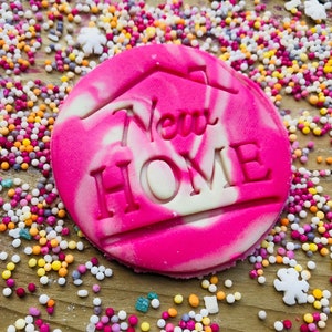 May include: A round, pink and white swirled cookie with the words "New Home" cut out. The cookie is surrounded by colorful sprinkles and small white flower-shaped candies. The cookie has a house outline above the words.