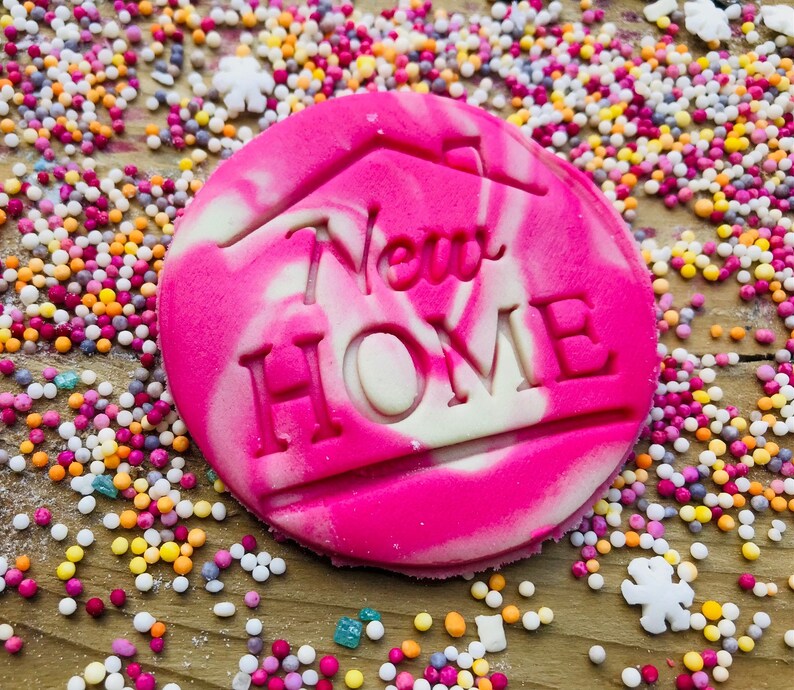 May include: A round, pink and white swirled cookie with the words "New Home" cut out. The cookie is surrounded by colorful sprinkles and small white flower-shaped candies. The cookie has a house outline above the words.