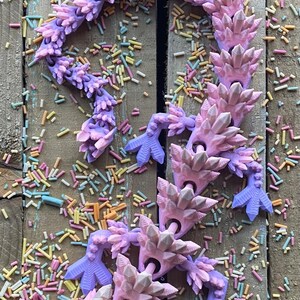 May include: A pink and purple crystal-shaped articulated dragon toy with a white crystal head. The dragon is lying on a wooden surface with colorful sprinkles scattered around it.