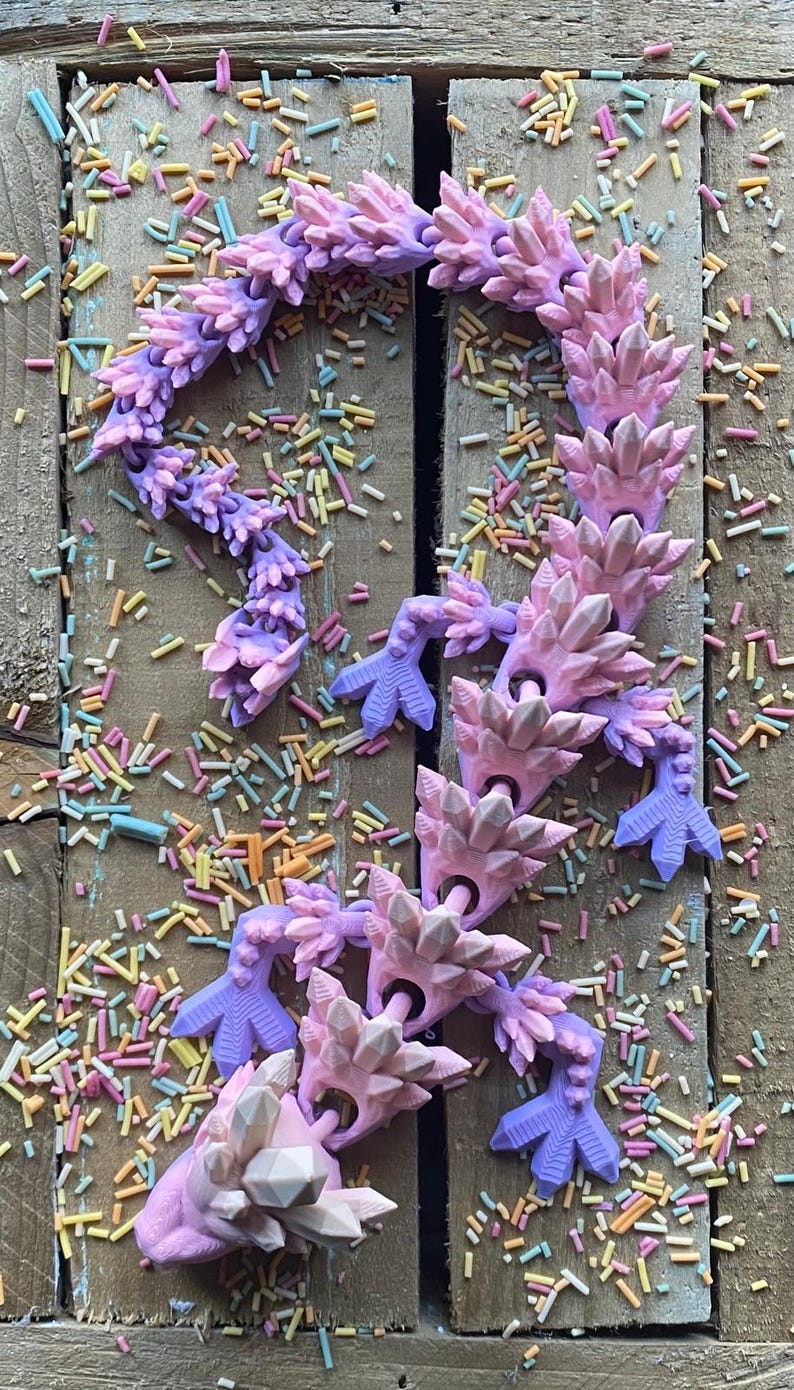 May include: A pink and purple crystal-shaped articulated dragon toy with a white crystal head. The dragon is lying on a wooden surface with colorful sprinkles scattered around it.