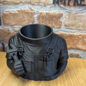 May include: A black planter shaped like a mechanic's torso, holding a wrench. The planter has a hollow head and is designed to hold plants. The mechanic's outfit includes pockets and overall straps, creating a unique decorative piece.