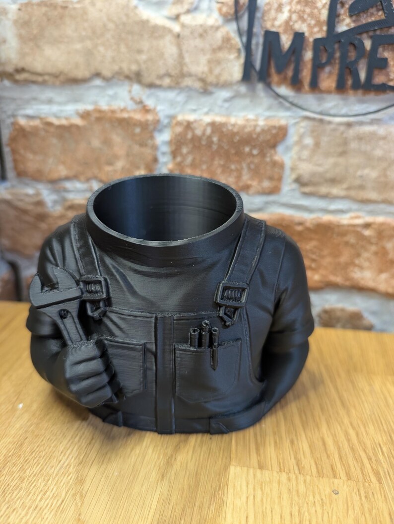 May include: A black planter shaped like a mechanic's torso, holding a wrench. The planter has a hollow head and is designed to hold plants. The mechanic's outfit includes pockets and overall straps, creating a unique decorative piece.